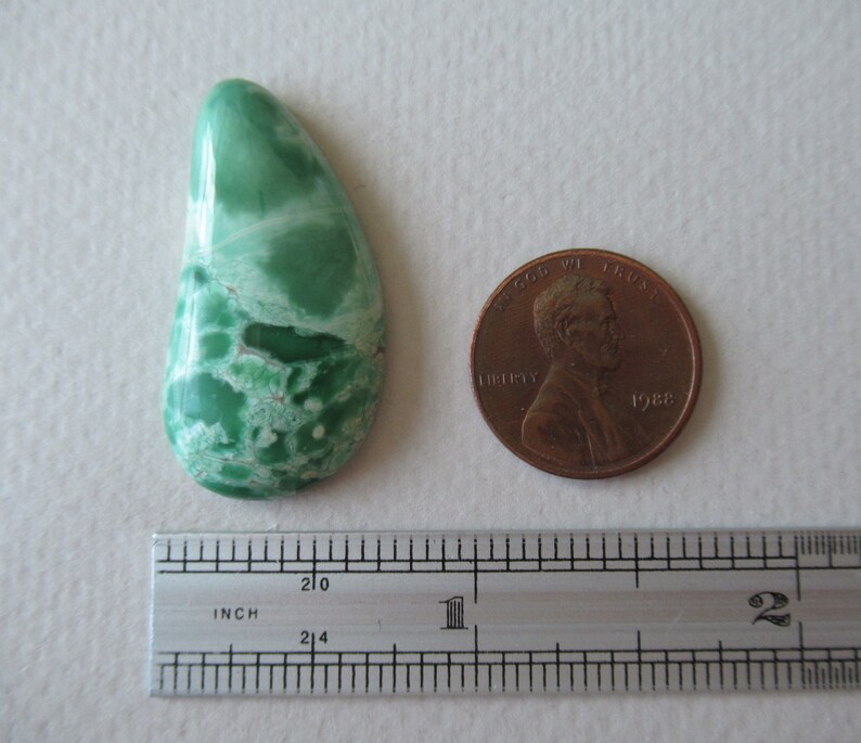 May include: A teardrop-shaped, polished gemstone with a vibrant green hue and white marbling. A copper penny and a ruler are next to the stone for size comparison. The stone's surface has a glossy finish, and the color variation creates a unique pattern.