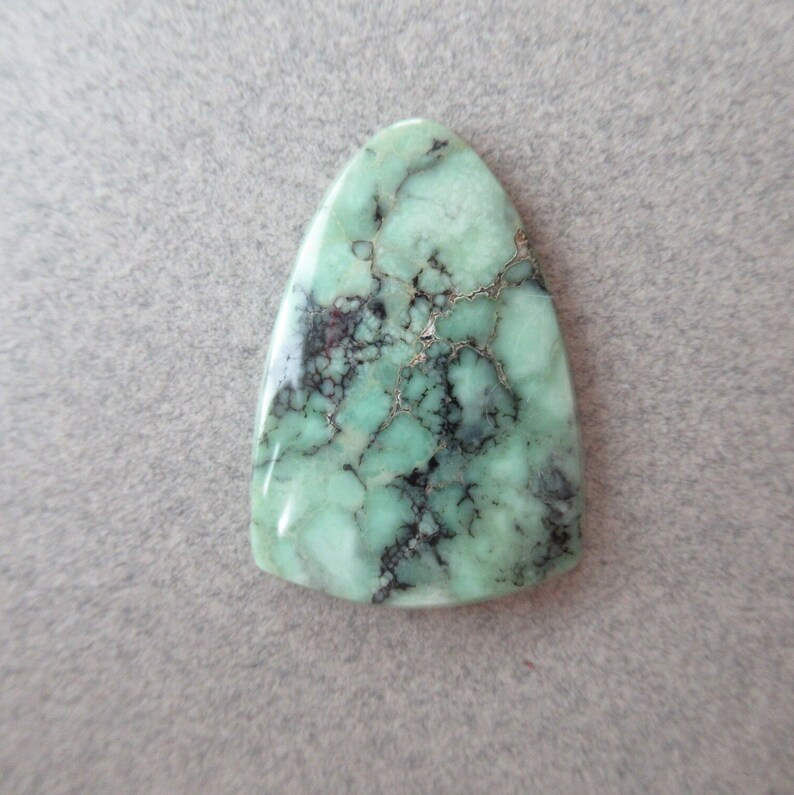 May include: A green and black gemstone cabochon with a teardrop shape. The stone has a marbled pattern with black veins.