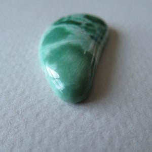 May include: A polished, teardrop-shaped gemstone with a smooth, glossy surface. The stone features a blend of light and dark green hues, with areas of white marbling. The gemstone is set against a neutral background, highlighting its natural beauty.