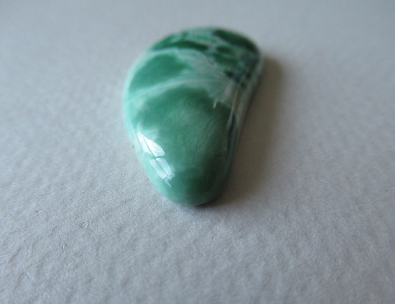 May include: A polished, teardrop-shaped gemstone with a smooth, glossy surface. The stone features a blend of light and dark green hues, with areas of white marbling. The gemstone is set against a neutral background, highlighting its natural beauty.