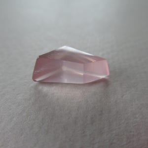 May include: A faceted, pale pink gemstone with a geometric form. The gem has a flat base and angled sides, creating a subtle glow. The stone is set against a neutral background.