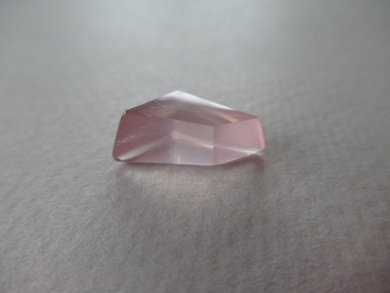 May include: A faceted, pale pink gemstone with a geometric form. The gem has a flat base and angled sides, creating a subtle glow. The stone is set against a neutral background.