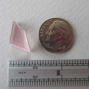 May include: A pale pink, geometric gemstone next to a British coin and a ruler. The gemstone has a faceted surface and a triangular shape. The ruler shows inch measurements. The coin features a profile of a man.