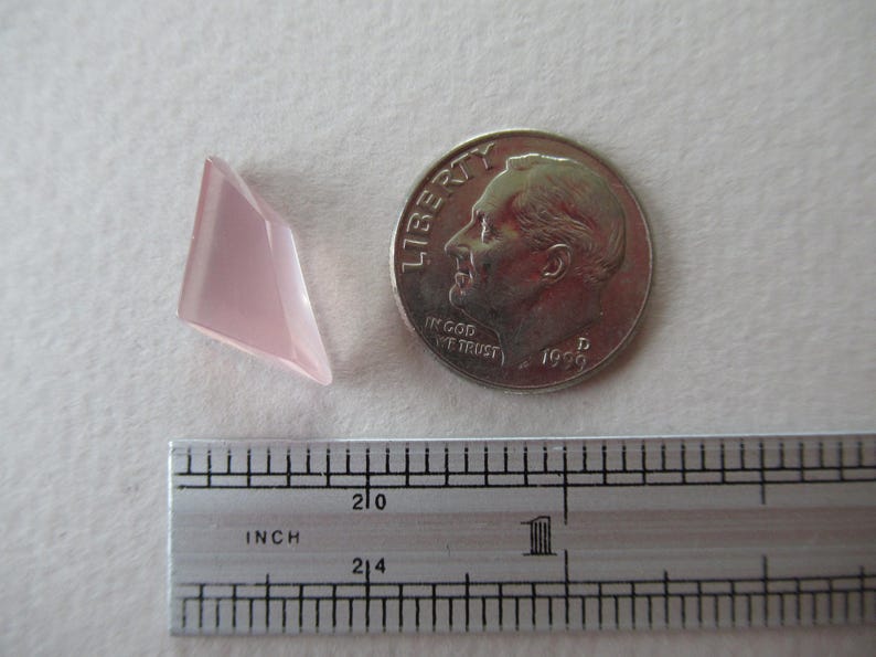 May include: A pale pink, geometric gemstone next to a British coin and a ruler. The gemstone has a faceted surface and a triangular shape. The ruler shows inch measurements. The coin features a profile of a man.