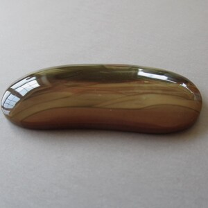 May include: A polished, oval-shaped stone with a smooth, glossy surface. The stone features a blend of brown, tan, and olive green hues, with a reflective quality that catches the light. The overall shape is elongated and rounded.