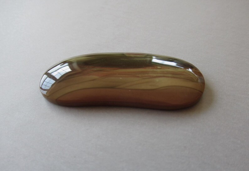 May include: A polished, oval-shaped stone with a smooth, glossy surface. The stone features a blend of brown, tan, and olive green hues, with a reflective quality that catches the light. The overall shape is elongated and rounded.