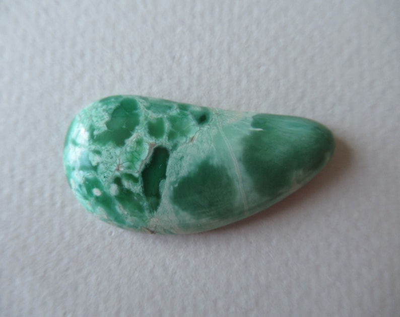 May include: A polished, teardrop-shaped gemstone with a mottled pattern of green and white. The stone's surface has a smooth, glossy finish, and the colors blend together in a natural, organic design. The gemstone is a unique, handcrafted item.