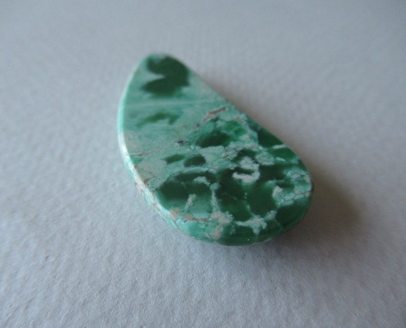 May include: A teardrop-shaped gemstone with a mottled pattern of light and dark green hues. The stone has a polished surface with a mix of textures and a light beige edge.