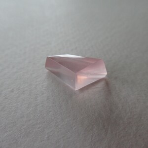 May include: A light pink, faceted gemstone with a geometric design. The translucent gem is set against a neutral background. The polished surfaces of the stone reflect light, creating highlights. The gem's shape is reminiscent of a cut crystal.