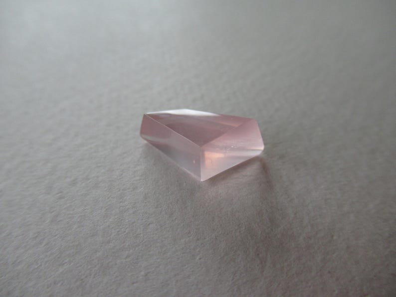 May include: A light pink, faceted gemstone with a geometric design. The translucent gem is set against a neutral background. The polished surfaces of the stone reflect light, creating highlights. The gem's shape is reminiscent of a cut crystal.