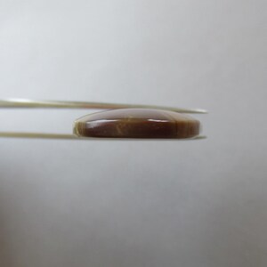 May include: A polished, oval-shaped gemstone, held by tweezers. The gem is a rich, warm brown color with subtle variations in tone. The surface appears smooth and reflective, suggesting a high-quality finish. The tweezers are silver-colored.
