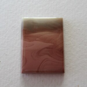 May include: A rectangular, polished gemstone with a layered appearance. The top portion is a light beige, transitioning to a brown and then a deep reddish-brown at the bottom. Swirling patterns create a natural, earthy aesthetic.