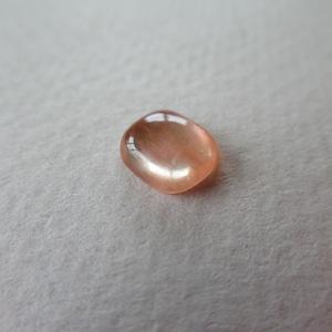 May include: A polished, oval-shaped gemstone with a peach-colored hue. The gem has a smooth, reflective surface, catching the light and displaying a subtle shimmer. The stone is set against a soft, off-white background.