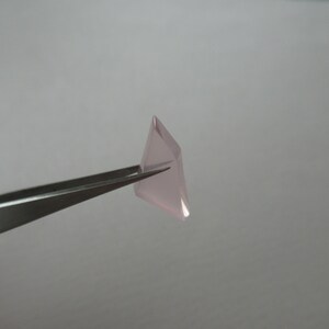 May include: A small, pale pink, faceted gemstone is held by silver tweezers against a plain, light grey background. The gemstone has a geometric shape with multiple flat surfaces.