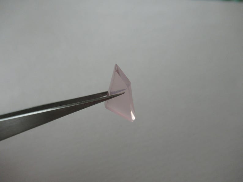 May include: A small, pale pink, faceted gemstone is held by silver tweezers against a plain, light grey background. The gemstone has a geometric shape with multiple flat surfaces.