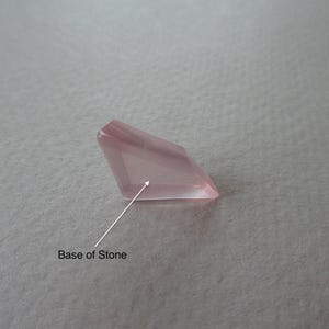 May include: A faceted, pale pink gemstone with a rectangular shape. The gem has a polished, translucent appearance, with visible facets catching the light. The base of the stone is indicated by an arrow. The background is a neutral, textured surface.