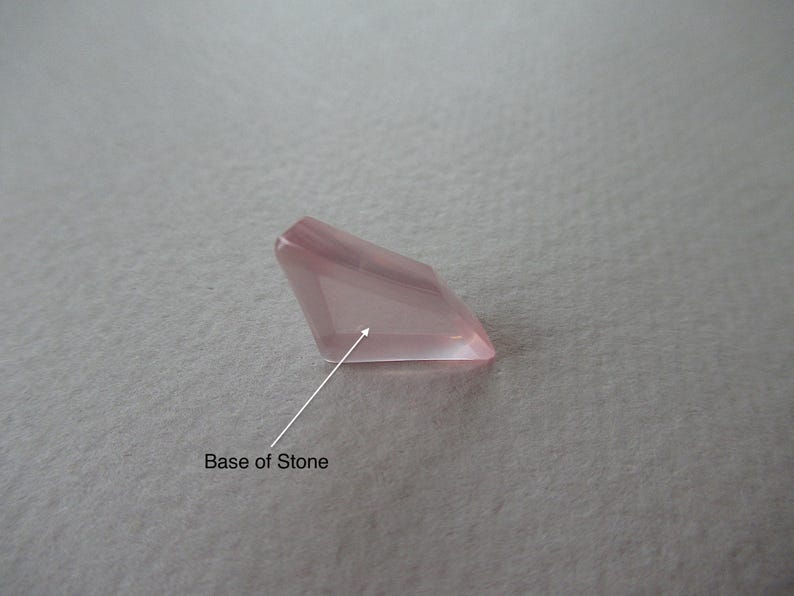 May include: A faceted, pale pink gemstone with a rectangular shape. The gem has a polished, translucent appearance, with visible facets catching the light. The base of the stone is indicated by an arrow. The background is a neutral, textured surface.