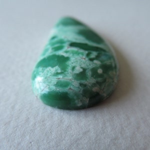 May include: A teardrop-shaped gemstone with a vibrant green hue, marbled with lighter green and white patterns. The polished surface reflects light, showcasing the natural variations and textures of the stone. The gemstone is a focal point, likely for jewelry or decorative purposes.