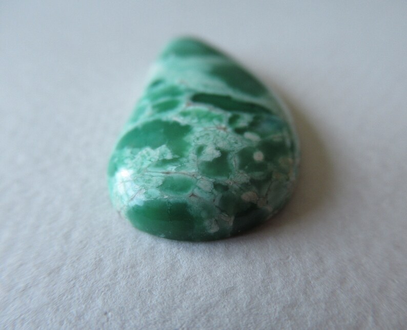 May include: A teardrop-shaped gemstone with a vibrant green hue, marbled with lighter green and white patterns. The polished surface reflects light, showcasing the natural variations and textures of the stone. The gemstone is a focal point, likely for jewelry or decorative purposes.