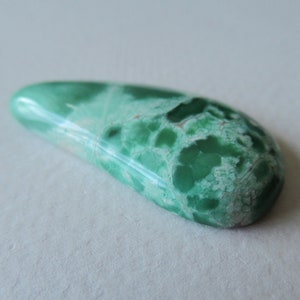 May include: A polished, elongated gemstone with a vibrant green hue, featuring a mottled pattern of lighter green and white. The stone has a smooth, glossy surface and a rounded shape, ideal for jewelry or collection.