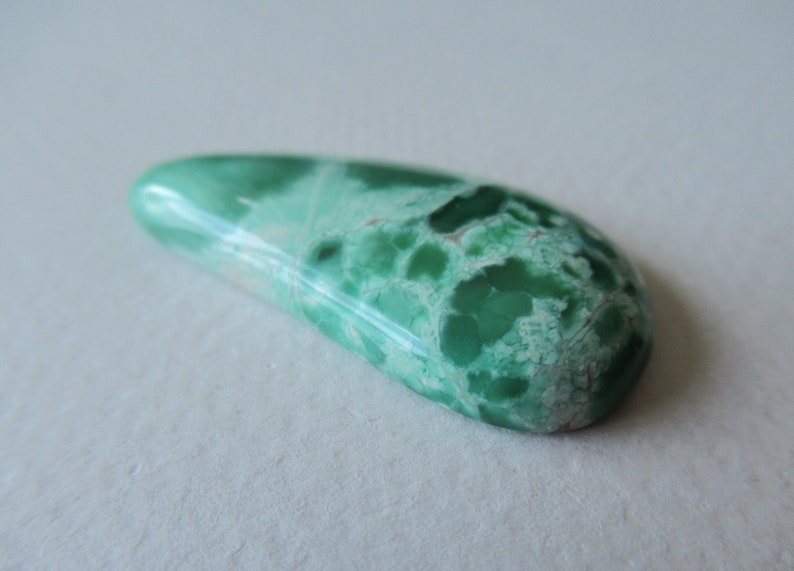 May include: A polished, elongated gemstone with a vibrant green hue, featuring a mottled pattern of lighter green and white. The stone has a smooth, glossy surface and a rounded shape, ideal for jewelry or collection.