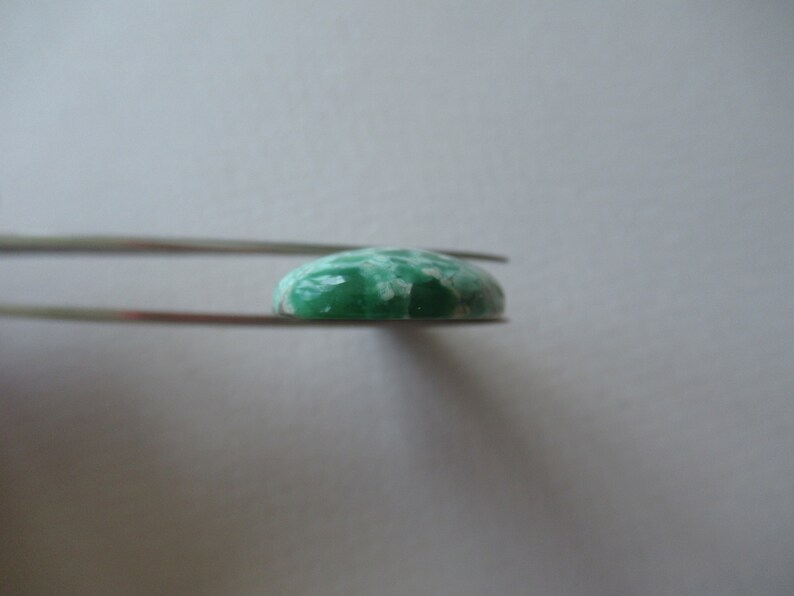 May include: A green and white gemstone held by tweezers. The gemstone has a rounded, oval shape with a polished surface. The color is a mix of light green and white, with a marbled appearance. The tweezers are silver.