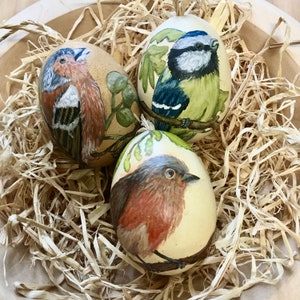 Op de afbeelding: Drie handgeschilderde eieren met verschillende vogelontwerpen. Eén ei heeft een blauwe vogel met groene bladeren, een ander heeft een bruine vogel met groene bladeren en het laatste ei heeft een rode vogel op een witte achtergrond.