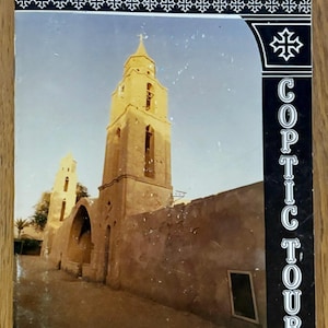 May include: A book cover featuring a photograph of the Anba Bishoi Monastery. The image shows a tall, beige-colored bell tower and a building with arched doorways. The text "(2) Anba Bishoi Monastery" is at the top, with "Coptic Tours" on the right side.