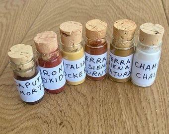May include: Six small glass bottles with cork stoppers, each containing a different colored powder. The bottles are labeled with handwritten text, including "Caput Mort", "Iron Oxide", and "Terra Sienna". The powders range in color from dark brown to white.