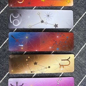 May include: Five colorful bookmarks with zodiac signs and constellations. The bookmarks are red, silver, blue, gold, and purple. Each bookmark features a different zodiac sign and constellation.