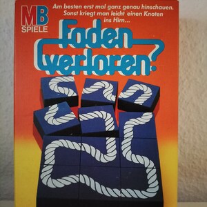 Könnte beinhalten: Eine Spielschachtel mit einem leuchtend orangenen Hintergrund und blauem Text, der "Faden verloren?" lautet. Das Spiel ist für Kinder ab 8 Jahren und für 1 oder mehr Spieler geeignet. Die Schachtel zeigt ein blaues und weißes Seilrätsel.