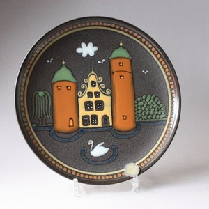 May include: A brown ceramic plate with a painted scene of a yellow house with two orange towers, a white swan, and green trees. The plate has a brown and gold border.