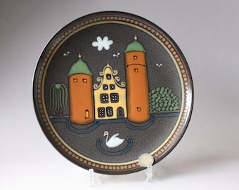 May include: A brown ceramic plate with a painted scene of a yellow house with two orange towers, a white swan, and green trees. The plate has a brown and gold border.