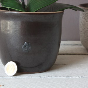 Könnte beinhalten: Ein dunkelbrauner Keramiktopf mit einer Münze auf dem Tisch davor.