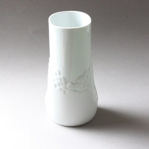 May include: A white ceramic vase with a raised floral design. The vase has a cylindrical shape with a slightly flared base.