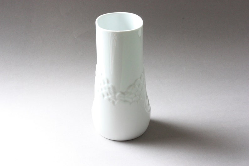 May include: A white ceramic vase with a raised floral design. The vase has a cylindrical shape with a slightly flared base.