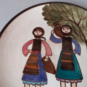 May include: A ceramic plate with a hand-painted design of two women in traditional clothing standing under a tree. The women are wearing brown dresses with colorful accents and carrying baskets. The tree is green with a brown trunk.