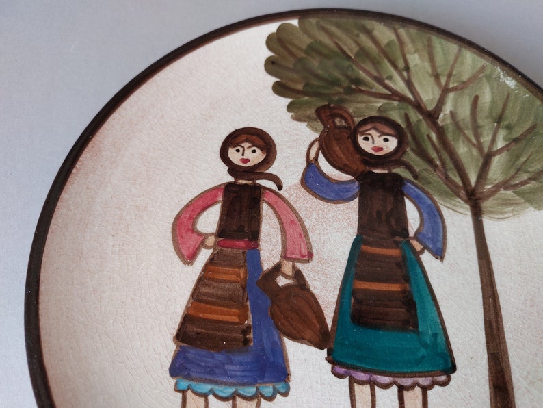 May include: A ceramic plate with a hand-painted design of two women in traditional clothing standing under a tree. The women are wearing brown dresses with colorful accents and carrying baskets. The tree is green with a brown trunk.
