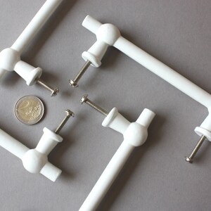 Op de afbeelding: Witte kastknoppen met een T-vormig ontwerp, met een centrale bol en twee loodrechte staven. Elke knop heeft een schroef voor montage. Een euromunt is inbegrepen ter referentie. Op een grijze achtergrond.
