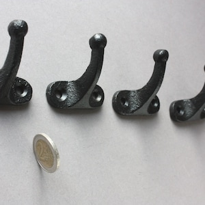 Könnte beinhalten: Fünf schwarze Metallhaken mit abgerundeten Köpfen, die an einer Wand montiert sind. Die Haken sind in einer Reihe angeordnet, mit einer Münze im Vordergrund.