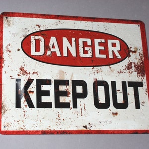 Könnte beinhalten: Ein weißes Metallschild mit roten und schwarzen Buchstaben. Das Schild lautet "DANGER KEEP OUT". Das Schild ist rostig und hat einen abgenutzten Look.