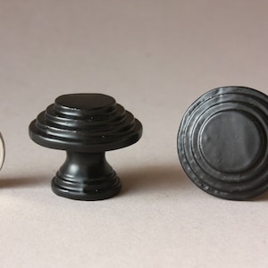 May include: Two black cabinet knobs. One knob is round with concentric circles. The other knob is a mushroom shape with a stepped top.
