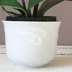 May include: White ceramic planter with a swirl design. The planter is empty and has a black foam insert at the top.