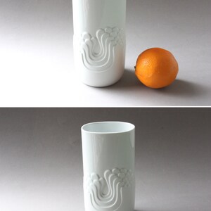 May include: White ceramic vase with a raised design of stylized waves. The vase is cylindrical and has a smooth, glossy finish.