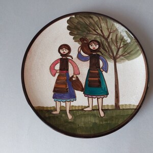 May include: A ceramic plate with a hand-painted design of two women in traditional clothing standing in front of a tree. The women are holding jugs and wearing colorful skirts and blouses.