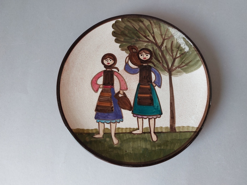 May include: A ceramic plate with a hand-painted design of two women in traditional clothing standing in front of a tree. The women are holding jugs and wearing colorful skirts and blouses.