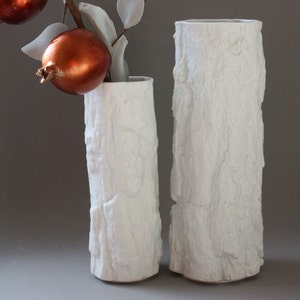 May include: Two white ceramic vases with a tree bark texture. The vases are cylindrical and have a smooth, white finish.