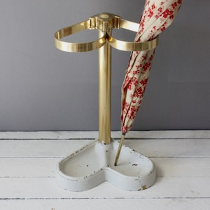 May include: A vintage umbrella stand with a white base and a brass-colored central pole and rings. An umbrella with a floral pattern in red and cream is placed in the stand. The stand has a heart-shaped base.