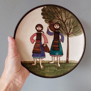 May include: A round ceramic plate with a painted scene of two women in traditional clothing standing in front of a tree. The women are holding a pitcher and a basket. The plate has a brown rim.