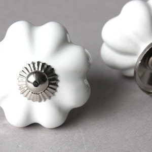 Peut inclure: Deux boutons de cabinet en céramique blanche avec un design cannelé et une base en métal argenté. Les boutons ont un bord festonné décoratif.